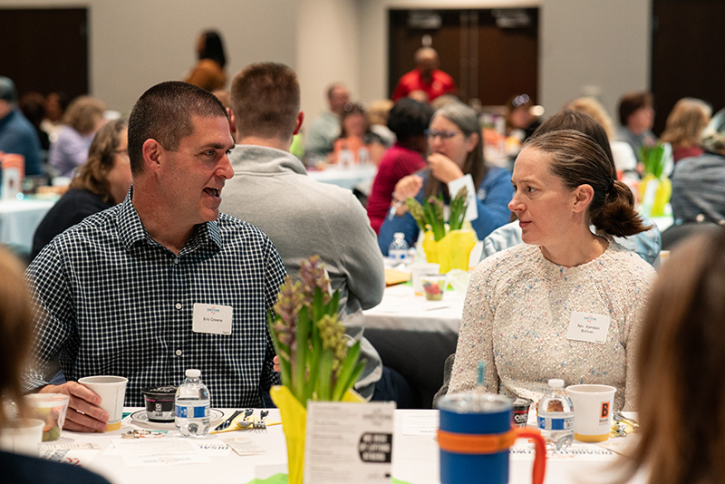 Candid photo of two breakfast event attendees visiting at their table, with other guests in the background.