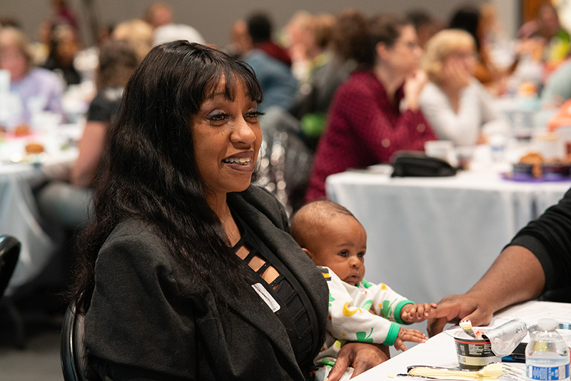 Candid photo of a breakfast event attendee holding a baby, with other guests in the background.