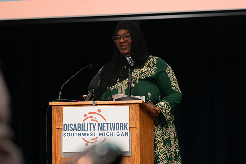 Speaker at a podium with microphones. The front of the podium holds a Disability Network sign.