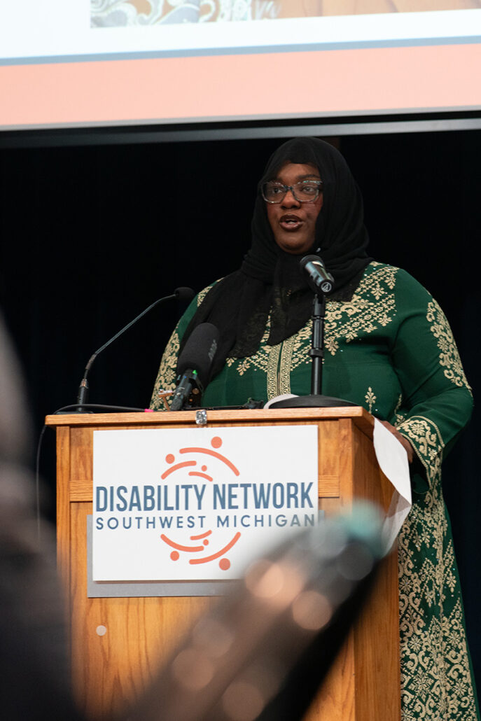Young, Black woman speaking at a podium with microphones. The front of the podium holds a Disability Network sign.