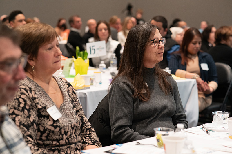 Candid photo of two breakfast event attendees watching the presentation, with other guests in the background.