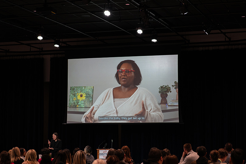 A video being shared on a large projector screen; image on the video is a young Black woman speaking about her personal experiences as a person with disabilities.