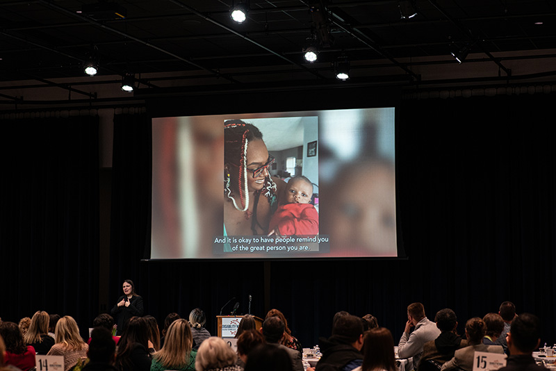 A video being shared on a large projector screen; image on the video is a young Black woman holding her baby.