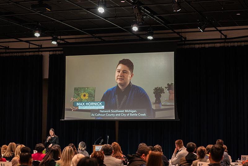 A video being shared on a large projector screen; image on the video is of a white Disability Network staff member being interviewed.
