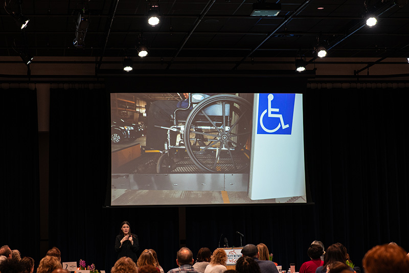 A video being shared on a large projector screen; image on the video is a person using a wheelchair which is being lifted into an accessible vehicle.