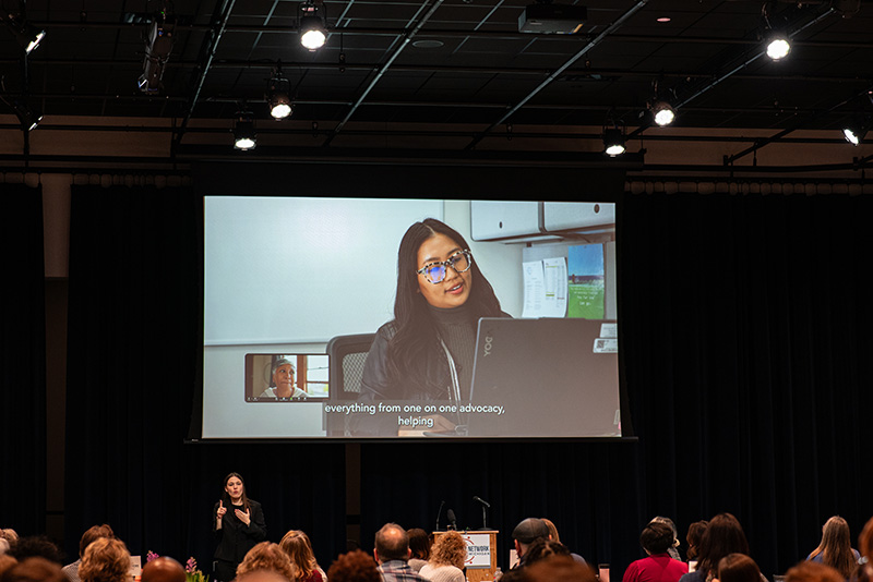 A video being shared on a large projector screen; image on the video is of a Burmese Disability Network staff member meeting virtually with a customer.