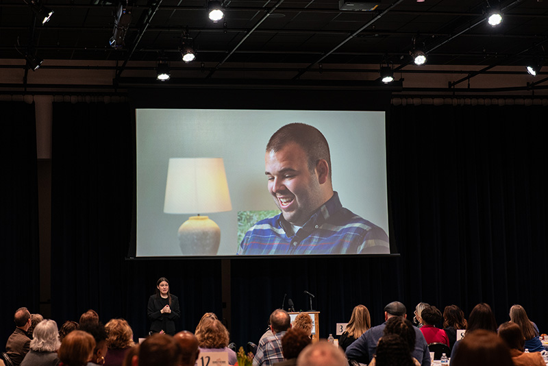 A video being shared on a large projector screen; image on the video is of a young, white, male Disability Network customer member being interviewed.