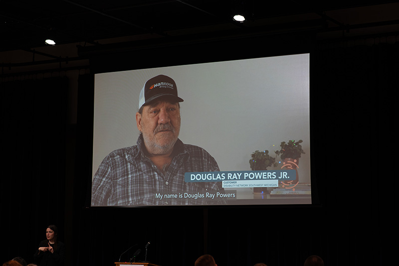 A video being shared on a large projector screen; image on the video is of a white, middle-aged, male Disability Network customer member being interviewed.