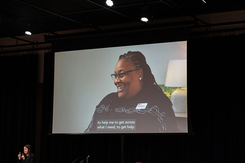 A video being shared on a large projector screen; image on the video is of a Black, middle-aged, female Michigan Works staff member being interviewed.
