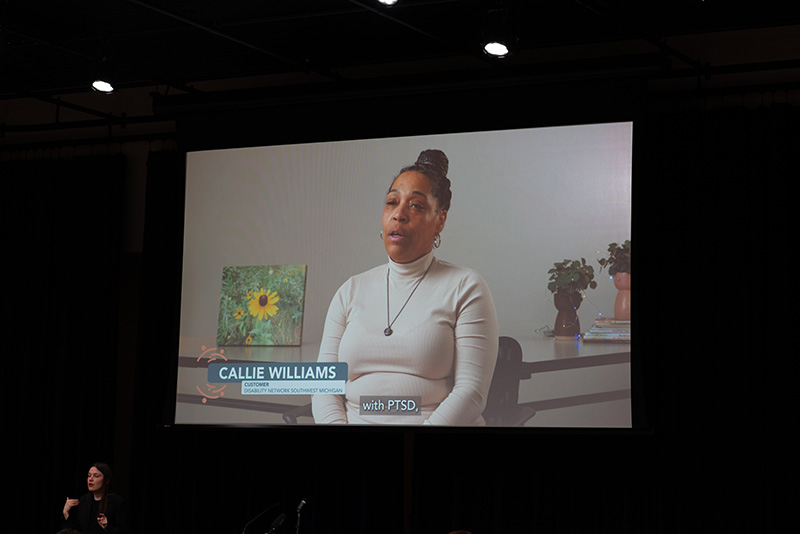 A video being shared on a large projector screen; image on the video is of a Black, middle-aged, female Disability Network customer member being interviewed.