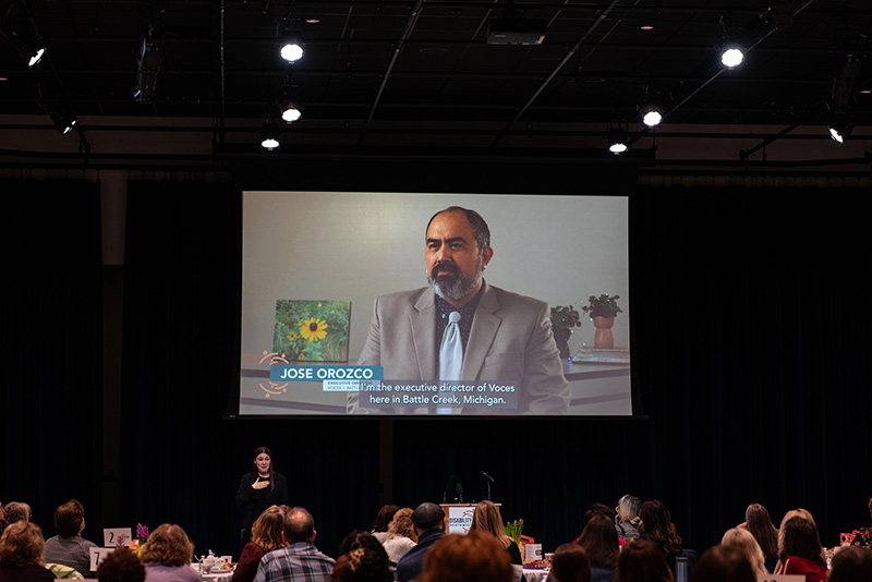 A video being shared on a large projector screen; image on the video is of a Latino, middle-aged, male VOCES staff member being interviewed.