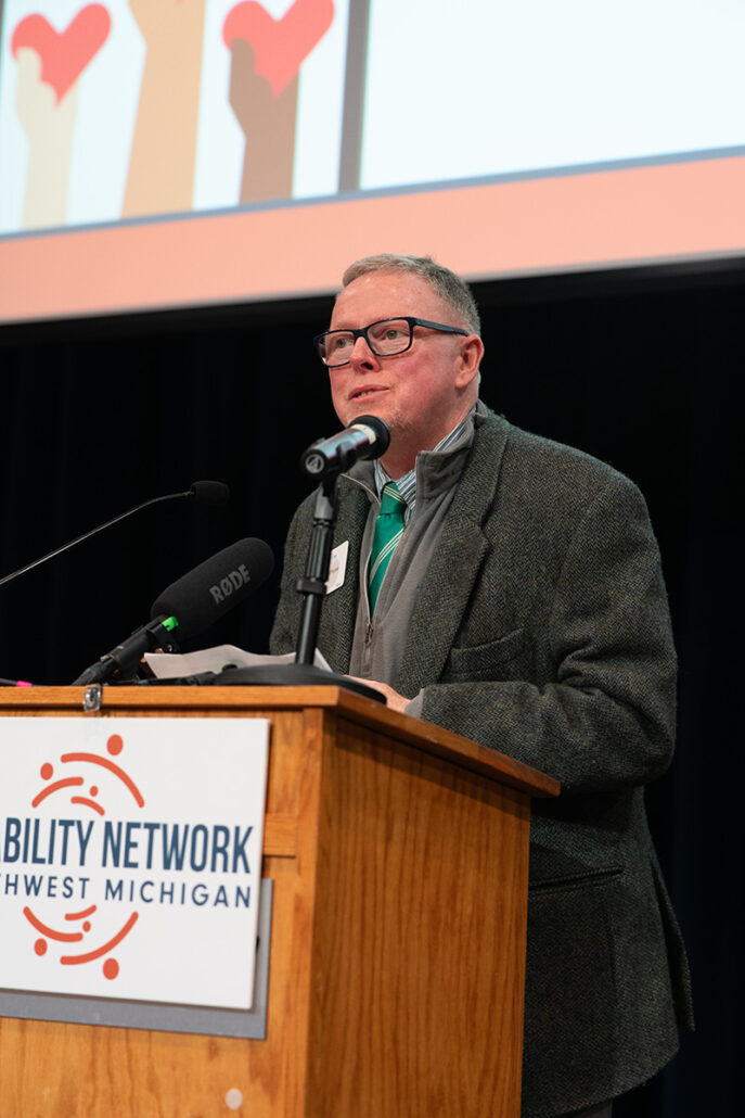 Middle-aged white man speaking at a podium with microphones. The front of the podium holds a Disability Network sign.