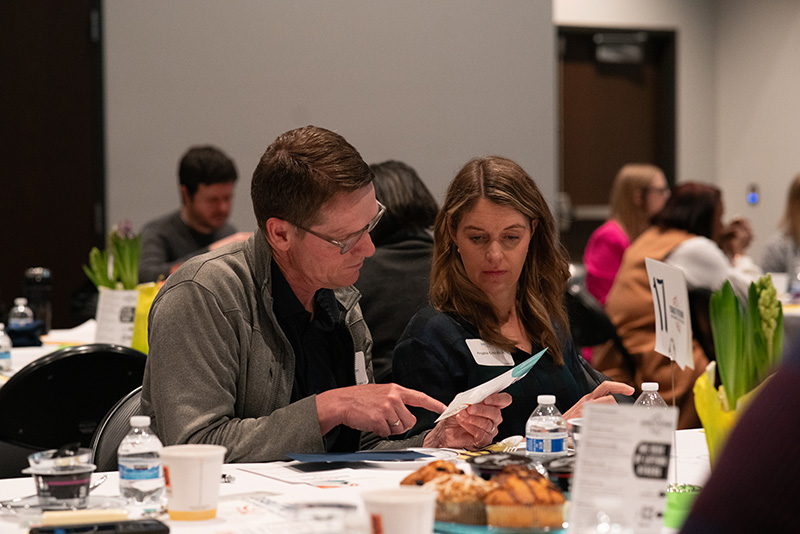 Candid photo of two breakfast event attendees at their table, looking at pledge cards and other information.