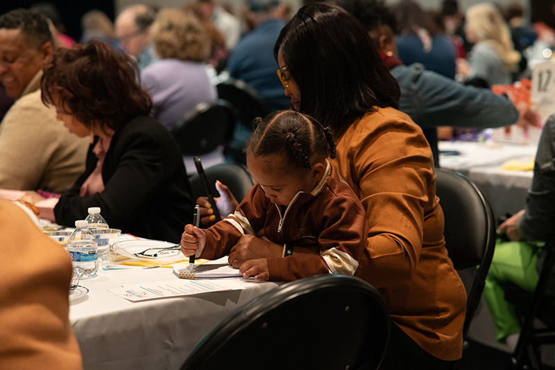 Candid photo of a breakfast event attendee seated at a table and holding a young child who is coloring. 