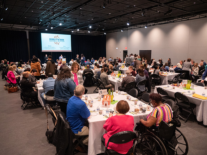 Candid photo of room full of tables and event guests. Disability Network logo is displayed on a large projector screen at the front of the room.