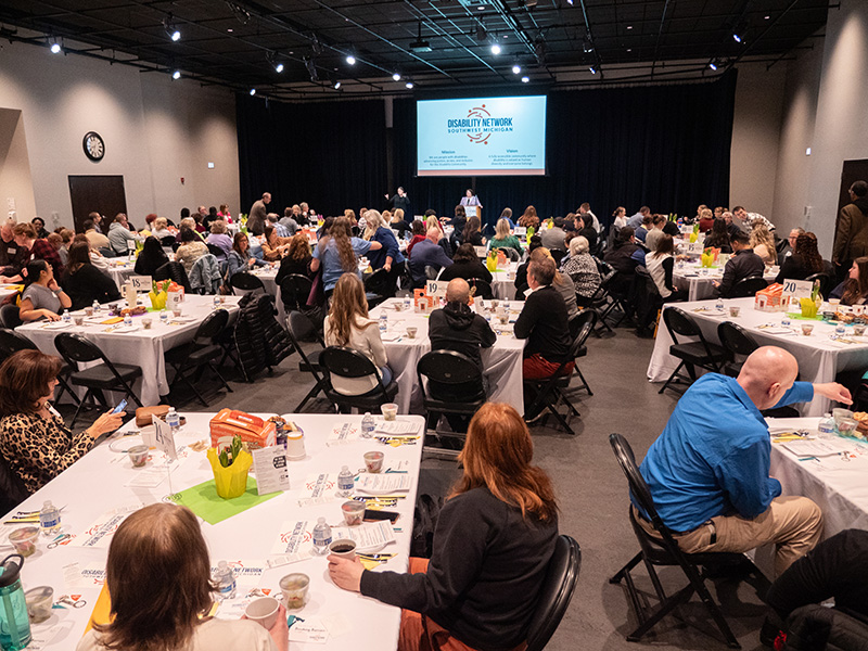 Candid photo of room full of tables and event guests. Disability Network logo is displayed on a large projector screen at the front of the room.